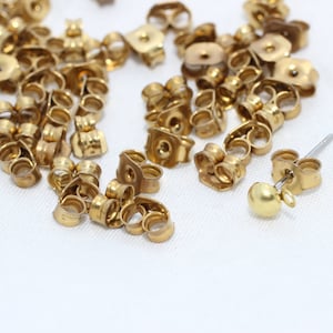 Peut inclure: Une collection de fermoirs de boucles d'oreilles dorés et une seule boucle d'oreille avec une boule dorée. Les fermoirs sont de différentes formes, dont papillon et disque. Les articles sont dispersés sur une surface blanche.