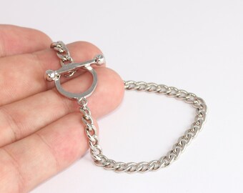May include: A silver-colored chain bracelet with a toggle clasp. The bracelet features a curb chain design and a circular clasp with a bar closure. The jewelry is displayed on a white background.