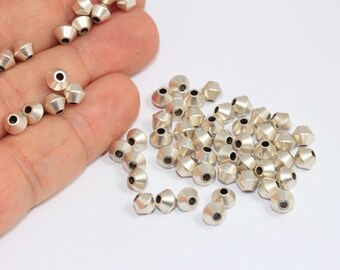 May include: Silver-colored metal beads with a faceted design and a hole through the center. The beads are scattered on a white surface, with some held in a hand. These beads are suitable for jewelry making and crafts.