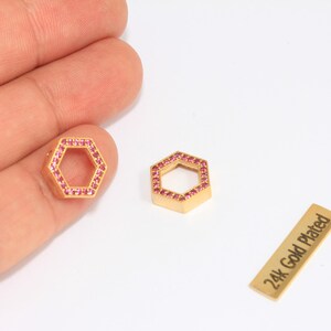 May include: Two gold-plated, hexagonal charms with pink gemstones. One charm is held by a hand. A small gold rectangle with the text "24k Gold Plated" is also present. The charms are likely for jewelry making.