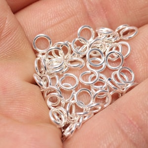 May include: A close-up of a hand holding a collection of small, silver jump rings. The rings are circular, with a small gap for attaching to other jewellery components. The image is well-lit, showing the metallic sheen of the rings.