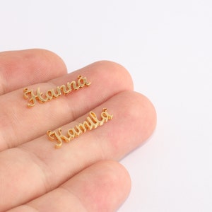 Puede incluir: Dos colgantes dorados con nombres, uno con la inscripción "Hanna" y el otro "Kamila". Los colgantes están en una fuente cursiva y parecen ser de metal. El fondo es blanco.