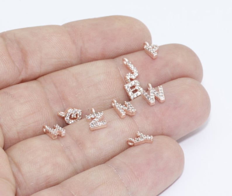 May include: A collection of rose gold letter charms, each encrusted with small, clear crystals. The letters include 'B', 'W', 'N', 'Z', and a heart shape. These charms are designed for jewelry making and personalization.