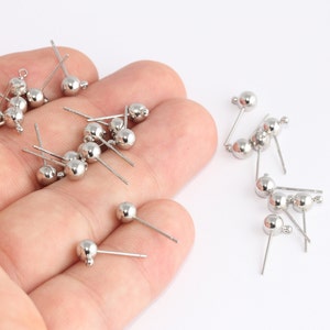 May include: A collection of silver-colored ball stud earrings with thin posts. The earrings have a small loop at the end of the post. The earrings are scattered on a white surface.