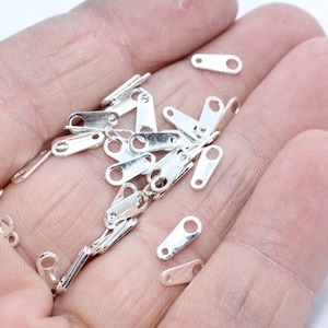 May include: A selection of small, shiny silver-coloured metal jewellery findings. The findings are rectangular with a hole at one end and a loop at the other. They are scattered across a light background.