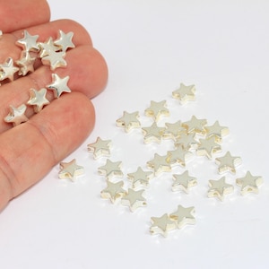 K&ouml;nnte beinhalten: Eine Sammlung kleiner, metallisch silberfarbener sternf&ouml;rmiger Perlen. Die Perlen haben eine glatte, reflektierende Oberfl&auml;che und ein f&uuml;nfzackiges Design. Einige der Sternperlen sind auf einer schlichten wei&szlig;en Oberfl&auml;che verstreut.