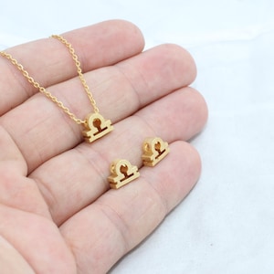 Può includere: Una collana e orecchini abbinati con il simbolo zodiacale della Bilancia color oro. La collana presenta una catena delicata e gli orecchini e il ciondolo hanno un design minimalista. I gioielli sono esposti su sfondo bianco.