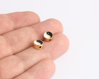 May include: Two gold-colored beads with a yin-yang design. Each bead features a black and white enamel yin-yang symbol. The beads have a cylindrical shape with a small hole for stringing. These beads are ideal for jewelry making.