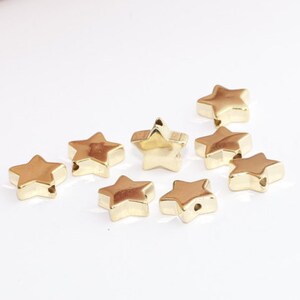 Könnte beinhalten: Eine Sammlung kleiner, goldfarbener sternförmiger Perlen. Jede Perle hat eine glatte, metallische Oberfläche und ein Loch zum Auffädeln. Die Perlen sind auf einer weißen Oberfläche verstreut, ideal für Schmuckherstellung oder Basteln.