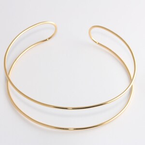 Può includere: Una collana dorata a girocollo con un design a doppio strato. La collana presenta una finitura liscia e lucida e un'estetica minimalista. Le due fasce parallele si incurvano per creare un look elegante e moderno. La collana è progettata per essere indossata intorno al collo.