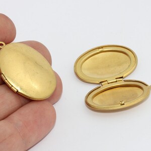 May include: Two gold-colored oval lockets, one closed and one open, displayed against a white background. The open locket reveals an interior space for a photo or keepsake. The closed locket is held in a hand.