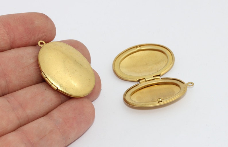 May include: Two gold-colored oval lockets, one closed and one open, displayed against a white background. The open locket reveals an interior space for a photo or keepsake. The closed locket is held in a hand.