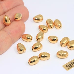 Op de afbeelding: Een verzameling goud vergulde, druppelvormige kralen. De kralen zijn glanzend goudkleurig en hebben een klein gaatje aan de bovenkant om te rijgen. Een klein rechthoekig label leest "24k Gold Plated".