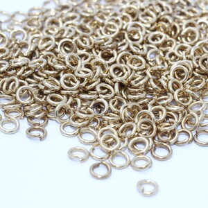 Op de afbeelding: Een stapel kleine, goudkleurige verbindingsringen, verspreid over een wit oppervlak. De ringen zijn cirkelvormig met een kleine opening, wat suggereert dat ze bedoeld zijn voor het maken van sieraden of knutselen. De belichting benadrukt de metaalglans.