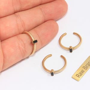 May include: Gold-colored rings with a black rectangular stone. One ring is on a finger, and two others are displayed separately. A small gold rectangle with the text "Raw Brass" is also present. Jewelry.