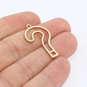 Op de afbeelding: Een goudkleurige metalen vraagtekenhanger. De hanger is een holle omtrek van een vraagteken, met een kleine lus aan de bovenkant om op te hangen. Het metaal heeft een matte afwerking. De hanger wordt in een hand gehouden.