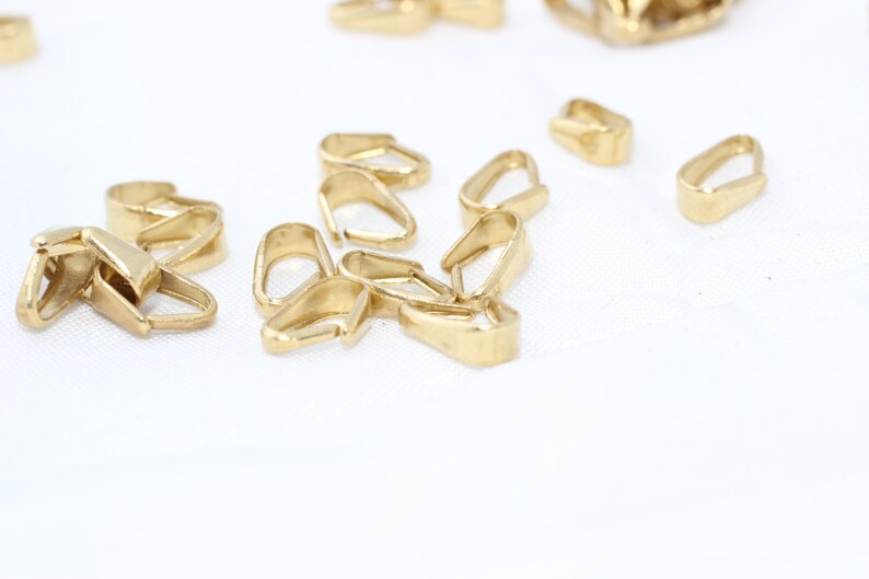 Puede incluir: Una colecci&oacute;n de peque&ntilde;os hallazgos de joyer&iacute;a dorados, incluidos conectores de fianza. Los hallazgos tienen varias formas, con un acabado liso y pulido. Est&aacute;n dispersos sobre una superficie blanca, lo que sugiere un entorno de artesan&iacute;a o joyer&iacute;a.