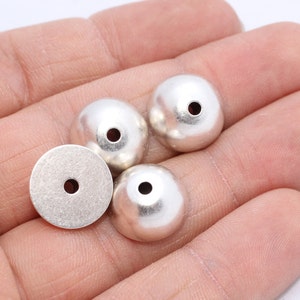 May include: A collection of silver-colored metal beads with a matte finish. The beads are round with a flattened top and a hole drilled through the center. One flat, round disc with a hole is also present.