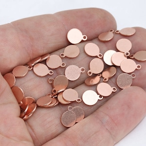 Puede incluir: Un conjunto de pequeños dijes redondos de metal color cobre, cada uno con un lazo en la parte superior. Estos dijes son ideales para la fabricación de joyas o proyectos de artesanía. Los dijes están dispersos sobre un fondo claro.