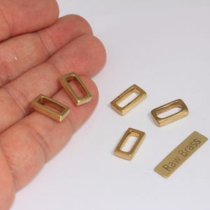 Puede incluir: Se muestran varios pequeños herrajes rectangulares de latón con esquinas redondeadas. También hay una pequeña etiqueta de latón con el texto "Raw Brass". Los herrajes son de color dorado cálido.