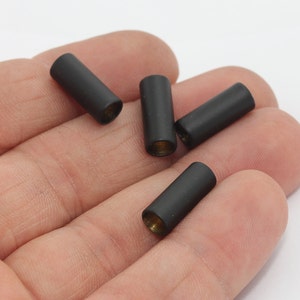 May include: Four matte black cylindrical beads with gold-colored interiors are displayed. The beads are evenly spaced and appear to be made of metal. The background is a plain white surface.