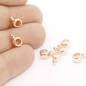 Pode incluir: Close-up de vários pequenos achados de joias douradas. Cada achado tem uma base circular com um pequeno laço que se estende da parte superior. Os achados estão espalhados em uma superfície branca, alguns sendo segurados em uma mão.