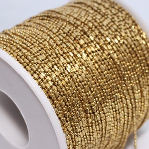 May include: A spool of gold-colored chain, tightly wound. The chain is made of small, rectangular links, creating a textured surface. The spool is white, providing a contrast to the chain's color. Ideal for jewelry making.
