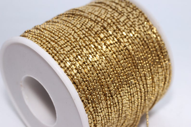 May include: A spool of gold-colored chain, tightly wound. The chain is made of small, rectangular links, creating a textured surface. The spool is white, providing a contrast to the chain's color. Ideal for jewelry making.