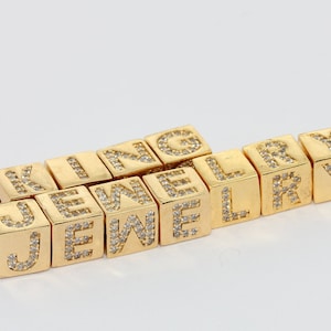 May include: Gold-colored cube beads spelling out the word "KING JEWELRY". Each cube has a letter encrusted with small, clear crystals. The beads are arranged on a white background.