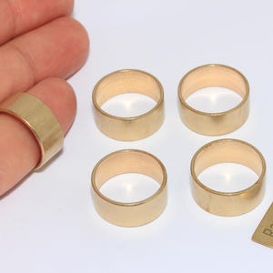 May include: Four raw brass rings and a small brass tag with the text "Raw Brass" are displayed on a white surface. One ring is on a finger. The rings are cylindrical and have a matte finish. The tag is rectangular.