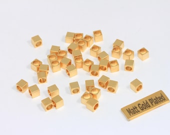 May include: A collection of small, gold-colored, square beads with a hole through the center, scattered on a white surface. A rectangular piece of gold-colored metal with the text "Matt Gold Plated" is also present. These beads are likely for jewelry making.