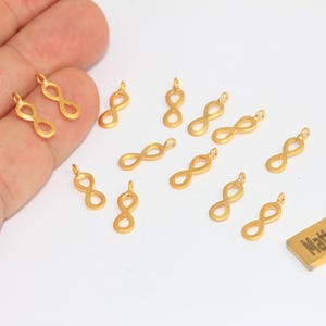 Könnte beinhalten: Goldfarbene Unendlichkeits-Anhänger. Die Anhänger haben die Form des Unendlichkeitszeichens, mit einer kleinen Schlaufe oben zum Befestigen an einer Kette oder einem anderen Schmuckbestandteil. Ein kleines rechteckiges Etikett trägt die Aufschrift "Matt Gold Plated".