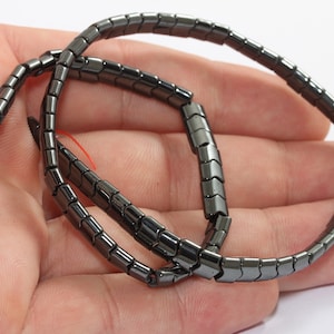 Può includere: Un filo di perline di ematite rettangolari grigio scuro. Le perline sono infilate su un filo rosso, formando una collana o un braccialetto flessibile. Le perline sono tenute nella mano di una persona.