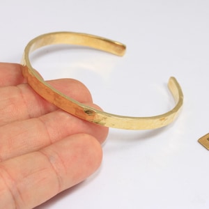 May include: A hammered brass cuff bracelet with an open design. The bracelet has a textured surface and a warm, golden color. A small brass tag with the text "Raw Brass" is also present.