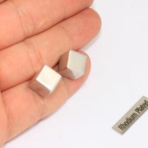 Op de afbeelding: Twee zilveren, kubusvormige manchetknopen worden in een hand gehouden. Er is ook een klein, rechthoekig label met de tekst "Rhodium Plated" aanwezig. De manchetknopen en het label staan op een witte achtergrond.