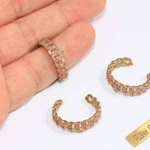 May include: Gold-toned brass jewelry components, including a ring and two open-ended cuffs, each adorned with small pink gemstones. A small brass tag with the text "Raw Brass" is also present.