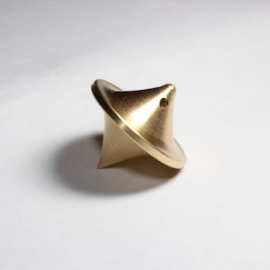 Op de afbeelding: Een massieve messing tol met een uniek, asymmetrisch ontwerp. De tol heeft een brede, gebogen basis en een puntige punt. Een klein gaatje is zichtbaar in de buurt van de bovenkant. Het metalen oppervlak reflecteert licht.