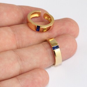 Puede incluir: Dos anillos dorados con una incrustación rectangular de piedra preciosa azul oscuro. Uno está en un dedo, el otro es abierto. Los anillos tienen una superficie pulida y reflectante.