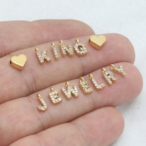 May include: Gold-colored jewelry charms in the shape of letters and hearts. The letters are encrusted with small, clear crystals. The charms are small and designed to be used in jewelry making.
