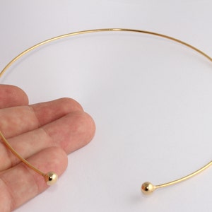 May include: A gold-colored choker necklace with a simple, elegant design. The necklace features a thin, curved band that widens at each end into a small, round, gold-colored bead. The necklace is shown against a white background.