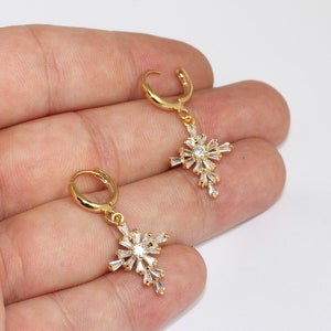 May include: Gold-colored hoop earrings with a cross pendant. Each cross is adorned with clear, sparkling crystals. The earrings are displayed against a white background.