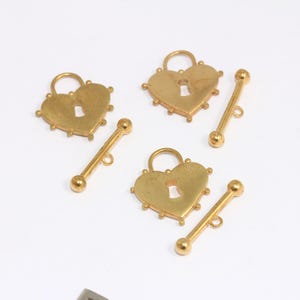 May include: Gold-colored heart-shaped lock pendants with keyholes and matching bar-shaped clasps. The heart-shaped locks have a textured surface and are surrounded by small, decorative studs. The clasps have a small loop at one end.