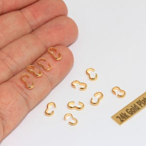 Può includere: Una collezione di piccoli coprinodo in metallo color oro con un design a forma di C. È presente una placca rettangolare color oro con il testo 24k Gold Plated stampato in nero. I componenti sono metallici, lucidi e di forma curva.