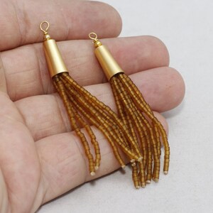 May include: Two gold-colored earrings with a cylindrical top and beaded fringe. The top portion is a matte gold, and the fringe is made of small, amber-colored beads. The earrings are shown against a neutral background.