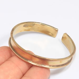 Könnte beinhalten: Ein goldfarbenes Armreif-Armband mit offenem Design. Das Armband hat eine rechteckige Form mit abgerundeten Kanten und einer glatten, polierten Oberfläche. Das Armband wird von einer Hand vor weißem Hintergrund gehalten.