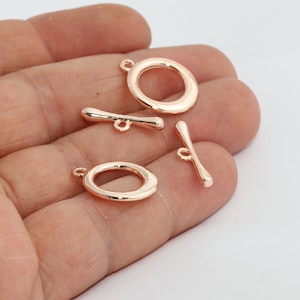 Könnte beinhalten: Roségoldene Knebelverschlüsse. Das Bild zeigt vier roségoldene Knebelverschlüsse. Zwei sind oval geformt mit einer Schlaufe, und zwei sind stangenförmig mit einer Schlaufe. Die Verschlüsse sind für die Schmuckherstellung.