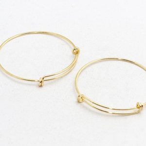 May include: Two gold-colored adjustable bangle bracelets. Each bracelet features a simple, circular design with a sliding mechanism for size adjustment. The bracelets are made of a shiny, metallic material.