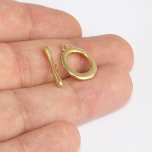 May include: A gold-colored toggle clasp set, featuring an oval ring and a bar. The clasp is designed for jewelry making and crafting. The set is shown against a neutral background, highlighting the metallic finish and simple design.