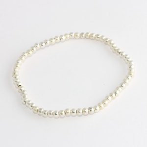 May include: A silver beaded bracelet. The bracelet is made of small, round, silver beads strung together on an elastic band. The beads are evenly spaced and create a simple, elegant design. The bracelet is shown on a white background.