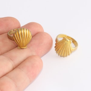 May include: Two gold-coloured adjustable rings, each featuring a detailed seashell design. The rings are made of a shiny metal and are set against a white background. The seashell design is the focal point of the rings.